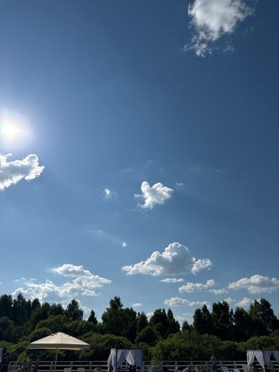 This image depicts a clear, bright day in Koloksha, Russia, under a vast blue sky dotted with fluffy white clouds. The sun shines intensely from the upper left corner, casting visible rays. In the foreground, a dense line of lush green trees forms a natural backdrop. Below the trees, a white railing delineates an area with lounge chairs and white canopies, suggesting a resort or recreational facility. A large, light-colored umbrella stands prominently near the center. Several people are visible in the lower portion of the image, some seated and others standing, enjoying the outdoor ambiance. The overall scene conveys a sense of a peaceful, sunny day in a natural setting within Koloksha.