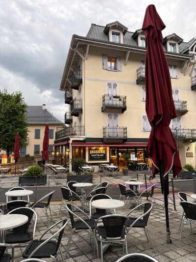 An exterior shot captures a cloudy day in Saint-Gervais-les-Bains, France, with a focus on a street-level restaurant. The building, painted a pale yellow, features multiple stories with balconies. A large, folded, dark red umbrella stands prominently in the foreground, obscuring part of the building and the street. The restaurant, named "Brasserie Mont-Blanc Saint-Gervais," has large glass windows and doors, with tables and chairs set up outside on a paved area. Several empty tables with chairs are arranged in front of the brasserie and extending into the foreground. To the left of the brasserie, a neighboring building is visible with a steeply pitched gray roof and dormer windows. A large tree with green foliage is positioned to the left of the frame. A single person, wearing a yellow shirt and blue shorts, is walking away from the camera on the right side of the scene, passing by the brasserie. The overall scene suggests a quiet, possibly early morning or late afternoon, atmosphere before or after peak dining hours, with the weather contributing to a somewhat subdued mood.