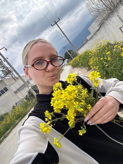 A blonde-haired female subject, with hair pulled back, wearing tortoiseshell-framed eyeglasses, is depicted in a close-up, self-taken photograph. She exhibits a slight pout and looks directly into the camera. She wears a black and white athletic jacket with a zippered front and a high collar. Her left hand holds a substantial cluster of freshly picked bright yellow wildflowers, consistent with Brassica species like mustard or rapeseed. Her fingernails are manicured, light-colored, and almond-shaped.

The scene is outdoors, situated along a paved or concrete pathway or road within the city of Kemalpaşa, Turkey. To the right of the subject, the ground is densely covered with the same yellow wildflowers, forming a field that extends into the midground. Overhead power lines are supported by multiple utility poles visible in the background. Industrial or commercial buildings with visible windows are present in the mid-distance on the left. In the further background, hills or mountains are visible beneath a partly cloudy sky, which features both bright and heavily clouded sections. Sparse, bare trees are also visible throughout the background.