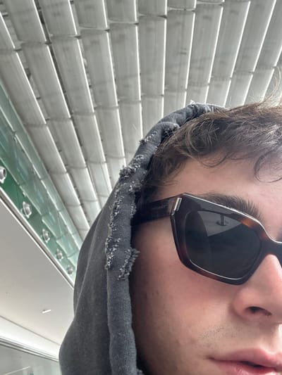 A selfie is taken indoors with a partial view of a person wearing dark sunglasses and a grey hooded sweatshirt. The person's face is close to the camera and partially angled. Blinds are visible in the background. The location is in Le Mesnil-Amelot, France.
