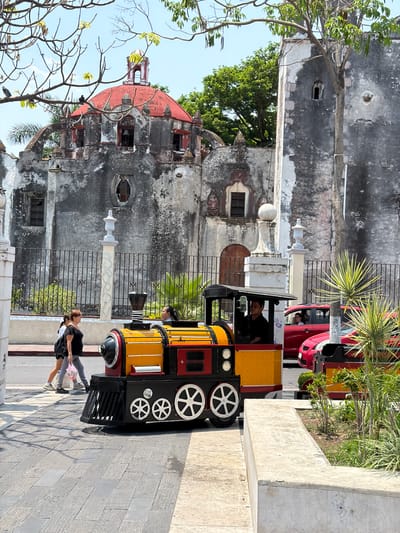 A ground-level shot of a colorful, toy-like locomotive in the foreground. The locomotive is primarily yellow and black with white wheels. A man is seen in the driver's seat. Two people, a taller person and shorter person, are seen walking near the front of the locomotive.

In the background, an aged stone building with a red dome is visible, likely a historical structure. A red car is parked in the background to the right. Some green foliage and a tree branch in the lower-left corner add natural elements to the scene. The setting is a paved area, and the image appears to be taken in daylight.

The location is Cuautla, Mexico.