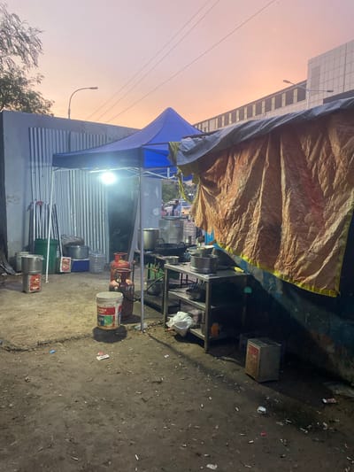 The image depicts an outdoor food stall setup in St. Thomas Mount-Pallavaram Cantonment Board, India, during twilight hours. A blue tent covers cooking equipment, including woks and gas cylinders, while a large orange tarp is draped over a makeshift structure to the right. The ground is uneven and dusty, littered with cigarette butts and debris. In the background, a modern building with a grid-like facade and streetlights suggest an urban setting. The sky displays a gradient of soft orange and pink hues, indicative of sunset or sunrise.