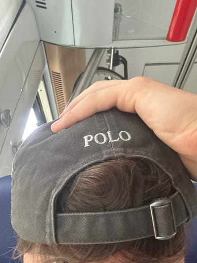 The image shows a person wearing a dark gray baseball cap. The cap has the word "POLO" embroidered in white on the back. The person's hand is resting on top of the cap. The background suggests an indoor setting, possibly a mode of transport such as a train, with muted lighting and visible ventilation grilles and reflective surfaces. There is no indication of the city of Salses-le-Château in France.