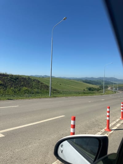 The image depicts a scenic view of a road in a rural mountainous region, likely near Altaiskoe, Russia. The road is asphalt, with a white line marking the lane. On the right side of the road, there are several red and white striped traffic cones. In the foreground, a car's side mirror is visible. Beyond the road, rolling green hills covered in grass and scattered trees extend into the distance, with more distant mountains forming the horizon under a clear blue sky. Several streetlights are visible along the roadside. The overall impression is of a well-maintained road passing through a picturesque natural landscape.