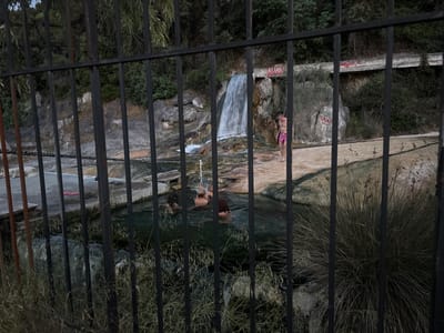 The image shows a fenced-off hot spring, likely located in the Municipal Unit of Lamia, Greece. A small waterfall cascades into the spring, which is occupied by several people bathing. One person is standing near the waterfall, partially disrobed. Clothing and footwear are visible on a nearby platform. Graffiti is scrawled on the concrete structure above the waterfall. The view is partially obstructed by the iron fence and vegetation.