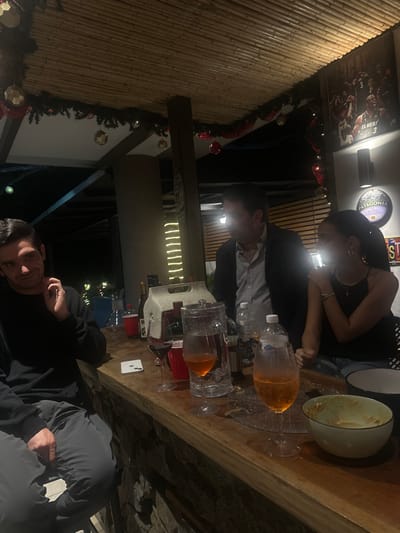 A social gathering is in progress at an outdoor venue decorated with Christmas garlands. A woman and a man are standing around a wooden table laden with pitchers of orange liquid and various beverages. An empty bowl sits nearby. Another man lies relaxed with a blanket on the lower portion of the frame looking at the camera. A framed basketball poster hangs on a wall in the background.