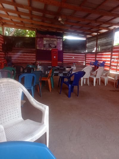 This image depicts an unoccupied, semi-enclosed structure in the city of Nara Unateze, Nigeria, likely serving as a gathering space. The structure features a corrugated metal roof supported by exposed wooden rafters and beams. The ground surface is unpaved, consisting of reddish-brown soil. One side of the enclosure is open, revealing green foliage, while another side is partially covered by horizontally striped red and white fabric or tarp material. A dark fabric backdrop is present at the rear.
In the foreground, an empty white plastic chair with a woven-patterned back is visible, along with a partial view of a blue plastic chair. Scattered across the unpaved floor are numerous empty plastic chairs of various colors including blue, green, orange, white, and black, arranged in loose rows and clusters.
At the rear of the space, a raised platform or stage is visible, covered with black and white checkered tiles. On this stage, a red, rectangular box-like object, possibly a podium or lectern, stands. Above the stage, affixed to the dark fabric backdrop, is a white rectangular sign displaying the text "KINGDOM RESTORATION REVIVAL MISSION" at the top, with further, less legible text below. A single white ceiling fan is mounted overhead near the center of the roof structure. No individuals are present in the scene.