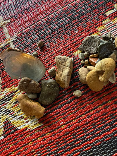 A close-up, high-angle shot shows a collection of natural objects on a woven mat with red, black, and yellow stripes. The mat's texture is intricate, with each strand appearing tightly twisted. Scattered across the mat are various stones, ranging in size, shape, and color from dark gray to light brown. Some stones are smooth and rounded, while others are more angular and rough. In addition to the stones, a large, iridescent mussel shell fragment is visible on the left side of the frame. The lighting suggests it is daytime, casting shadows that highlight the textures of the objects and the mat. The scene appears to be laid out for display or examination, possibly in an outdoor setting in Parapat, Indonesia, given the natural elements. No people are present in the image.