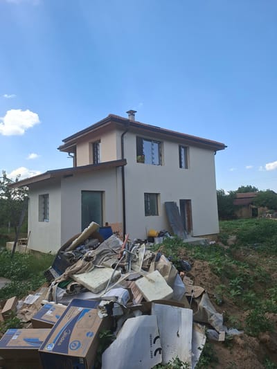 In the city of Dolna Malina, Bulgaria, a two-story house with light beige walls and a brown tile roof is under construction or renovation. The house has multiple windows, some with visible frames and glass, and a chimney on the roof. A man is visible looking out of an upper-story window. In front of the house, a large pile of construction debris, including cardboard boxes, foam insulation, pipes, and various other materials, is accumulated. The area surrounding the house is overgrown with green vegetation and appears to be a rural or semi-rural setting. The sky is blue with scattered white clouds.