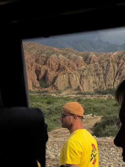 The image captures a man standing in the foreground, facing left, with his back partially towards the camera. He wears a yellow t-shirt with a graphic design on the back and a brown beanie. He has sunglasses on and appears to be looking out at a vast, rugged landscape. Behind him, a dry, rocky terrain with sparse green vegetation stretches towards imposing, multi-colored rock formations, characteristic of the canyons near Tong, Kyrgyzstan. The scene is framed by the interior of a vehicle, suggesting the man is either arriving at or departing from this location. The sky above is partly cloudy, with a muted, hazy appearance.