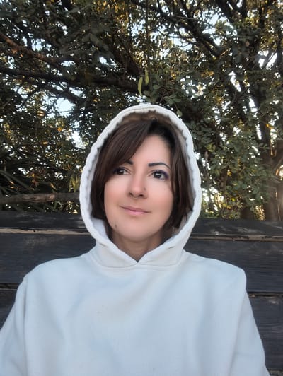 A medium close-up portrait shows an adult individual wearing a light-colored, off-white hooded sweatshirt with the hood up, positioned outdoors in Shaalvim, Israel. The individual has short, dark brown hair with bangs, brown eyes accented with purple eyeliner on the right eye and black on the left, and subtle glitter below the outer corner of both eyes. A small gold-toned hoop nose ring is present on the right nostril, and a small dark mole is visible on the lower left cheek. A silver-toned chain-link earring is visible on the left ear. The person displays a slight smile and is looking upward and to the viewer's left, away from the camera. Behind the individual, a dark wooden structure, appearing to be a bench or wall composed of horizontal planks, is visible from mid-torso level downwards. The upper background features a dense canopy of trees with dark green leaves and visible branches, through which small patches of light blue sky are discernible. The scene is illuminated by natural, dappled light.