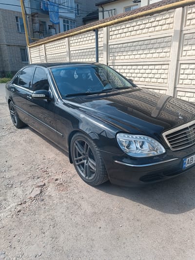 The image shows a black Mercedes-Benz S-Class sedan parked on a dirt road. The car is the primary subject, viewed from the front quarter. The background includes a building with grey brickwork and a yellow pipe running along the top of a concrete fence. The fence has a brick-like pattern. The scene suggests an outdoor setting, possibly in a residential or industrial area. The time of day appears to be daytime, with strong shadows cast by the car and the fence, indicating bright sunlight. The weather seems to be clear.
The car's headlights are illuminated. The license plate is visible, with the text "AB 4" and the Ukrainian flag symbol. The car's reflection of the concrete fence is visible on its hood and windshield. The ground beneath the car is composed of gravel and dirt, with some loose stones. There are no people visible in the image. The overall impression is of a static scene, with the car parked and stationary. The location context provided indicates Mohyliv-Podilskyi, Ukraine, which aligns with the visible license plate.