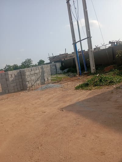 The image depicts an outdoor construction site in Tasha, Nigeria, under a clear sky. In the foreground, a dirt road shows tire tracks. To the left, a large stack of grey concrete blocks is arranged neatly. Behind the blocks, a red shipping container is partially visible. In the mid-ground, more stacks of concrete blocks are present, along with a pile of small grey gravel. Several tall utility poles with wires dominate the center and right side of the image. Beyond the poles, a long, grey wall with a darker top section is visible, and on top of the wall, there is barbed wire fencing. Further in the background, there are trees. The overall impression is of an active, albeit unrefined, construction area.
