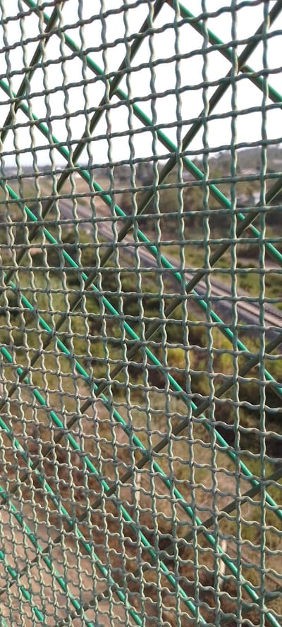 The image displays a close-up, high-angle shot looking through a green wire mesh fence, which occupies the majority of the foreground and is in sharp focus. The fence is constructed from corrugated or wavy wires, interwoven to form a grid of roughly square openings. The wires are a dull, possibly faded or dusty green color, with some areas appearing lighter or showing signs of wear.

Through the openings of the mesh, a heavily blurred outdoor background is visible. The soft focus of the background creates a sense of depth, emphasizing the fence as the primary subject. In the middle distance of the blurred background, several parallel lines are discernible, strongly suggesting railway tracks or train tracks stretching into the distance. These tracks appear to be on a slightly elevated or open terrain. Beyond the tracks, the landscape consists of indistinct patches of green, likely vegetation such as trees or grass, interspersed with lighter, brownish areas that could be dry ground or dirt. The sky or distant horizon is very bright, indicating it is daytime.

No specific activity is taking place; the image captures a static scene. The vantage point seems to be from an elevated position, possibly a bridge or an overlook, given the downward perspective on the tracks. There is no visible text in the image.