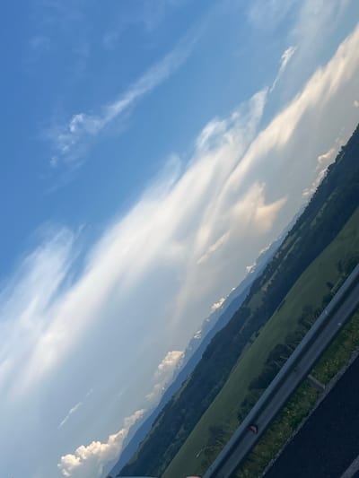 The image, taken in Vakarel, Bulgaria, shows a scenic landscape from a roadside perspective. The view is dominated by a vast blue sky filled with wispy white clouds, with a prominent band of elongated clouds stretching diagonally across the frame. Below the sky, a lush green hillside slopes downwards, dotted with dark green trees and patches of lighter green fields. In the distance, layers of mountains recede into the hazy horizon, with hints of snow on their peaks. A metal guardrail runs along the bottom right of the image, separating the road from the hillside. The road itself, in the foreground, appears to be dark asphalt, with some gravel and grass along its edge. The image is taken at an angle, giving a dynamic, tilted perspective of the landscape.