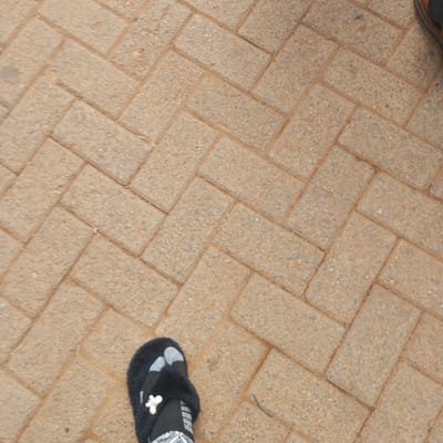 The image shows a section of a brick-paved area, likely a walkway or patio. The bricks are rectangular and laid in a herringbone pattern. The bricks are a light brown/tan color with a textured surface. In the bottom left corner, there is a foot wearing a black, fuzzy slipper with a white decorative flower. In the top right corner is part of another person’s shoe, possibly a brown shoe. The location is possibly Sheywe ward, Kenya, based on the prompt, but there are no indicators within the image itself to confirm this location.