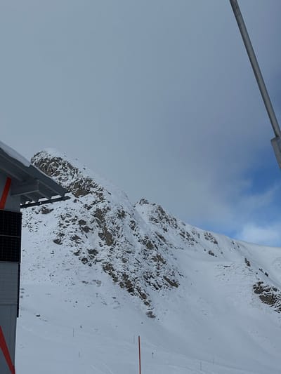 The image presents a snow-covered alpine environment in Bad Hofgastein, Austria. The scene is dominated by a rugged, snow-dusted mountain range under a partially overcast sky, transitioning from uniform grey clouds to a patch of blue with white clouds in the upper right. In the foreground, on the left, a portion of a building with a light grey facade is visible, featuring black rectangular solar panels and two vertical orange stripes. Its snow-covered roof extends horizontally. On the far right, a grey metallic pole, likely part of a ski lift system, ascends vertically. The mid-ground and lower sections display extensive snow-covered slopes, presumably ski pistes, marked with faint ski tracks and several thin, orange marker poles. No individuals are present.