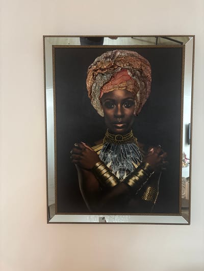 The image displays a framed portrait of a Black woman wearing a colorful headwrap and elaborate jewelry, including a large gold necklace with feather-like accents and gold armbands. She has her arms crossed, and her expression is neutral. The portrait is framed by a decorative border that appears to include mirrored sections and a beaded trim. The artwork is hanging on a plain, light-colored wall. The reflection in the mirrored frame shows parts of a room, including what appears to be furniture and possibly a television. The scene is indoors.