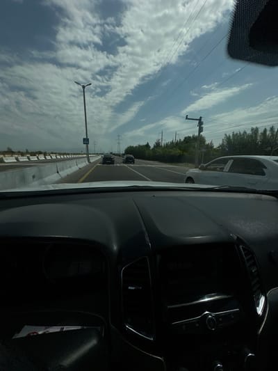 The image, taken from inside a vehicle, shows a highway scene with multiple lanes of traffic. The sky is partly cloudy, with patches of blue visible. Several cars are driving on the road, and power lines stretch across the landscape. On the left side of the road, a concrete barrier separates the lanes, and a pedestrian crossing sign is visible. In the distance, trees line the side of the road, and power transmission towers can be seen. The dashboard of the vehicle in the foreground is dark and features a digital display. The overall scene appears to be a typical highway environment.