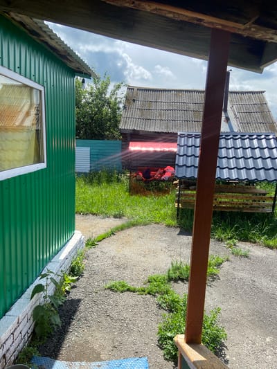 The image depicts an outdoor scene in Askarovo, Russia, under a cloudy sky. In the foreground, a green corrugated metal building is visible on the left, with a white-framed window. A path made of gravel and asphalt leads away from the building. To the right, a wooden swing set with a grey corrugated metal roof is present. On the swing, a person is lying down, partially obscured by red light filtering through a translucent barrier. Several plush toys are also on the swing. In the background, older wooden structures with grey corrugated roofs are visible. The area is covered with green grass and some weeds.