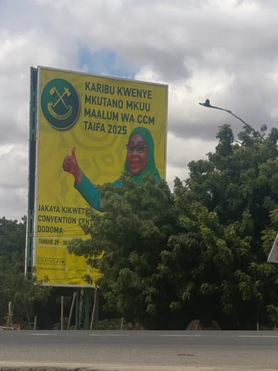 A large billboard is on the side of a road in Dodoma, Tanzania (latitude: -6.166, longitude: 35.784) with a picture of a woman wearing a green headscarf and glasses with her right thumb raised in the air. Text on the billboard reads "Karibu kwenye mkutano mkuu maalum wa CCM Taifa 2025" and "Jakaya Kikwete Convention Centre Dodoma Tarehe 29 - 30 M" along with various social media icons. The billboard is partially obscured by green foliage. The road is paved and there is a small piece of trash lying in the middle of it. The sky is overcast.