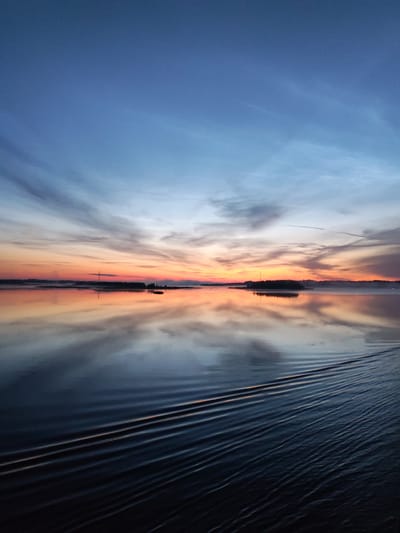 The image depicts a tranquil waterfront scene at sunrise or sunset, characterized by a serene body of water reflecting the colorful sky. The sky exhibits a gradient of colors, transitioning from a deep blue at the top to shades of orange, pink, and yellow near the horizon, suggesting the presence of clouds. In the distance, a silhouetted treeline runs along the horizon, punctuated by a prominent tower-like structure. The water's surface is largely calm, with clear reflections of the sky and the distant landscape. In the foreground, there are subtle ripples and wave patterns on the water, indicating movement, possibly from a boat or wind. The environment appears to be natural and unpopulated, with no people or explicit urban elements visible. The scene evokes a sense of peace and natural beauty. The location in Novodevich'e, Russia, is not visually identifiable from the provided image.