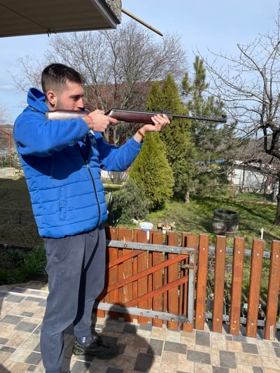 In a residential area of Zdravets, Bulgaria, a young adult male is depicted standing on a tiled patio, aiming a long-barreled air rifle. The male, observed in a profile view facing right, wears a blue hooded jacket over a blue puffer vest, dark blue sweatpants, and black athletic shoes. Colorful bracelets adorn both wrists. He holds the air rifle, which features a dark wooden stock and a black barrel, with both hands, supporting the buttstock against his right shoulder and aligning his head to sight down the barrel.

The immediate foreground consists of a brown and tan tiled patio. Directly behind the subject is a reddish-brown wooden fence with vertical slats, supported by a metallic frame, partially open as a gate. Beyond the fence, a grassy yard with several trees is visible; some trees are bare deciduous, while others are evergreen conifers. A wooden barrel is situated on the grass further into the yard. Distant structures, including parts of a white building and additional fencing, are present in the background under a clear, bright blue sky with sparse clouds.