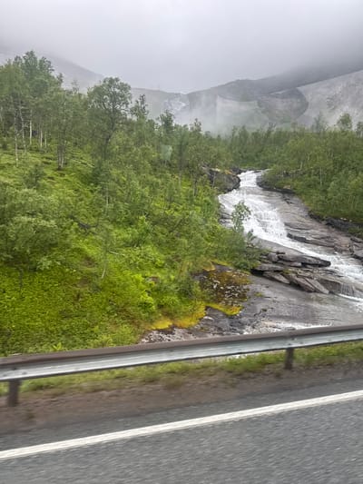 The image depicts a cascading waterfall flowing over dark, rocky terrain, bordered by lush green foliage and trees. In the background, misty mountains are visible under an overcast sky. In the foreground, a grey asphalt road with a white line runs horizontally, separated from the natural landscape by a silver guardrail. The scene is set in Mørsvikbotn, Norway, suggesting a natural, scenic environment. There are no people or animals visible in the image.