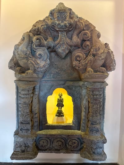 The image shows a stone niche in a wall. The niche is highly decorated with carvings featuring swirls and possibly figures. Inside the niche, illuminated by a warm light from behind, is a small, dark statue. The niche appears to be part of a larger architectural structure, possibly an ancient building. The wall surrounding the niche is plain and light-colored. Based on the prompt, the location of the image is in the city of Lalitpur, Nepal.