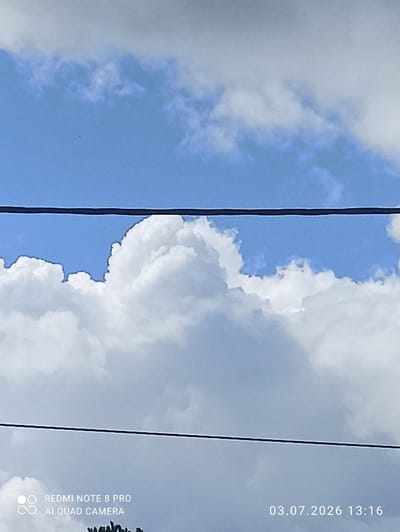 The image is a close-up of a cloudy sky. Two black wires stretch horizontally across the frame, dividing the sky into sections. The top section of the sky is a bright blue, with some wispy white clouds visible. The middle section is dominated by large, puffy white clouds that obscure much of the blue sky. The bottom section is a lighter gray, indicating more distant or less illuminated clouds. In the lower left corner, there is text that reads "REDMI NOTE 8 PRO AI QUAD CAMERA". In the lower right corner, there is text that reads "03.07.2026 13:16". The image does not show any people, buildings, or any indication of the city of Sovetskiy, Russia.