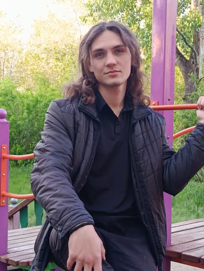 A young man with long, wavy brown hair is seated on a painted wooden structure outdoors, likely a playground in Lisakovsk, Kazakhstan. He is wearing a black polo shirt and a quilted black jacket. He is holding onto a horizontal bar attached to a purple support post with his right hand. The structure he is sitting on has a wooden bench seat and colorful posts and bars. The background consists of green foliage and trees, suggesting a park or recreational area. The overall setting appears to be a casual, outdoor environment.