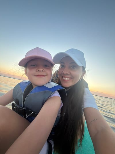 A woman and a young girl are taking a selfie on a body of water, possibly a lake or river, with a sunset visible in the background. Both are wearing life jackets, suggesting they are on a boat, kayak, or paddleboard. The girl is in the foreground, looking directly at the camera with a slight smile. She is wearing a pink baseball cap and a gray and blue life jacket over a purple shirt. The woman is to her right, also smiling and looking at the camera. She has long dark hair and is wearing a white baseball cap and a white t-shirt. Her left arm is extended, holding the camera. A faint tattoo of the word "balance" is visible on her forearm. The water is calm, and the sky is a pale blue, transitioning to orange and pink near the horizon. The location is Salaspils, Latvia, and the time appears to be during the golden hour of sunset.