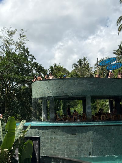 The image shows a multi-level structure with a pool in Tegal Suci, Indonesia. The structure has a green tiled exterior and overlooks a lush, green landscape with trees and vegetation. People are present on the upper levels of the structure, some appearing to be at a bar area with seating. The pool extends along the side of the structure. Blue decorative circular objects are visible in the background. The sky is overcast with clouds.