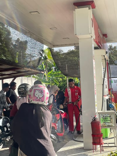 The image depicts a gas station scene in Glee Gogo, Indonesia. A person in a red uniform, presumably an attendant, is seen dispensing fuel into a motorcycle. Several individuals are waiting in line on their motorcycles, with some wearing helmets. One helmet is notably decorated with a pink Hello Kitty design. In the background, lush green foliage and trees are visible, suggesting an outdoor setting. A red fire extinguisher is mounted on the gas pump. Some text is visible on a sign behind the gas pump, although it is difficult to decipher clearly. The lighting suggests it is daytime, with bright sunlight. The overall atmosphere appears to be a typical daily transaction at a local gas station.