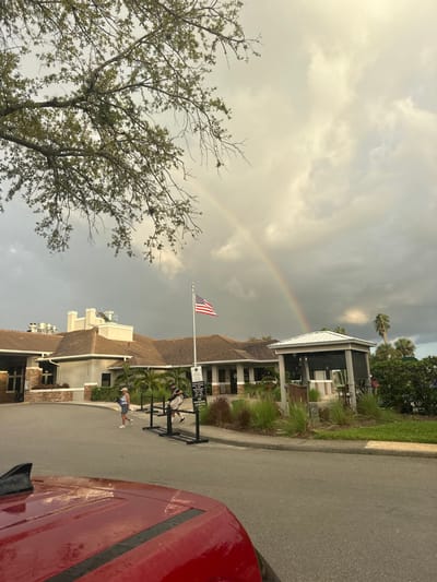 This image depicts an outdoor scene at a facility in Estero, United States, under a partly cloudy sky with a visible rainbow. In the immediate foreground, the red hood of a vehicle is visible, indicating the perspective. A paved asphalt driveway curves from the left towards the right, leading past a large, multi-sectioned building with a brown tiled roof and light-colored walls, consistent with a clubhouse or community center. An American flag is flown from a flagpole positioned centrally in front of this building. To the right of the flagpole, a small, open-sided gazebo-like structure with a light-colored roof stands beside the driveway. Two individuals, both appearing to be in golf attire and carrying golf bags, are walking from left to right on the paved area in front of the building. Black, portable stanchions or barriers are arranged near the flagpole. The sky above is largely overcast with grey clouds, but a distinct rainbow brightly arcs across the upper right portion of the frame. Vegetation includes a large tree with sparse foliage in the upper left, numerous palm trees, and various landscaped shrubs and grasses around the structures.