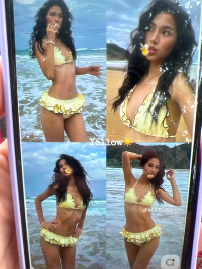 The image is a photograph of a person wearing a yellow bikini with ruffled edges, posing in the ocean. The person has dark hair and a flower in their mouth. The scene appears to be a beach or coastal area with waves and a sandy shore in the background. The image is presented on a phone screen, with reflections and light glare visible. The city of Kara-Dıykan, Kyrgyzstan is not depicted in this image.