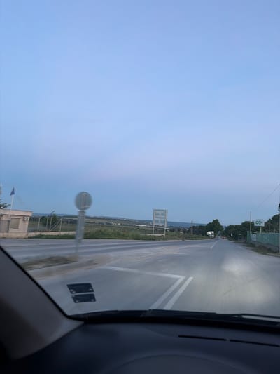 The image depicts a view from the perspective of someone inside a car, looking out through the windshield. The car is on a paved road, seemingly at an intersection or a point where the road curves. The sky is a pale blue, suggesting it might be early morning or late evening.
To the left, there is a low, light-colored building with a window. Adjacent to this building, flags are visible. A traffic signpost with a circular, gray sign is present in the middle ground, slightly out of focus. Beyond the road, there is a grassy area with some vegetation and a fence. In the distance, rolling hills are visible, with some scattered lights indicating a town or settlement.
To the right of the road, there is a green fence and trees lining the area. A large sign with the letters "GC" in green on a white background is prominently displayed. Further down the road, a white truck is visible, along with a few other vehicles and streetlights.
The image is captured from inside a vehicle, with the dashboard and windshield frame visible in the foreground, indicating a point-of-view shot. There are two rectangular stickers with multiple circular markings on the lower part of the windshield. The overall scene suggests a road passing through a rural or semi-rural area near a settlement.