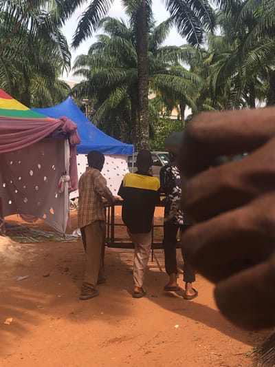 The scene is set in Nteje, Nigeria, with palm trees visible in the background. There are three individuals present, though one is heavily obscured by what appears to be a hand held close to the camera. The other two individuals stand near a makeshift structure covered with colorful fabric and a dotted white sheet. One person wears a plaid shirt and khaki pants, while the other wears a black shirt with a yellow stripe and light-colored pants. The ground is reddish-brown dirt.