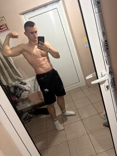 The image captures a young adult male in an interior room within the city of Pitovo, Bulgaria, via a mirror selfie. The male, exhibiting a muscular physique with visible abdominal muscles and short, buzzed hair, stands shirtless, wearing black athletic shorts with a red waistband and "NIKE AIR MAX" branding, along with white socks featuring two black stripes. He is flexing his right bicep while holding a black smartphone in his left hand, positioned to photograph his reflection. The room features a tiled floor composed of light-colored square tiles and light beige walls. A white PVC door or door frame with a white handle is prominently visible on the right, bearing the text "VIVA ALUMINIUM SYSTEMS" and "AVIV." To the left of the male's reflection, a white radiator is partially obscured by draped clothing, including a garment with a white snowflake motif. A light-colored bag and a black electrical cable are also present on the floor near the radiator. A framed item with partially obscured text is visible on the far left wall, and a small, colorful drawing is affixed to the wall on the right.