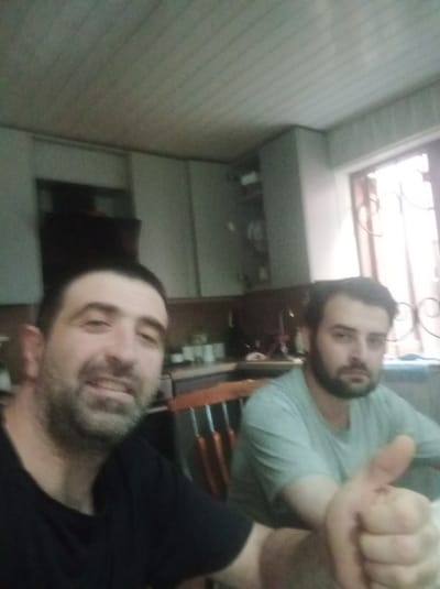 Two men are pictured indoors, possibly in a home or restaurant setting, in Poti, Georgia. The man in the foreground on the left is smiling and wearing a black t-shirt. The man on the right is giving a thumbs-up and wearing a light blue t-shirt. The background shows kitchen cabinets and a window with a decorative grille. The lighting suggests it could be daytime, but it's difficult to determine the exact time. The overall mood appears casual and friendly. There is no visible text in the image.