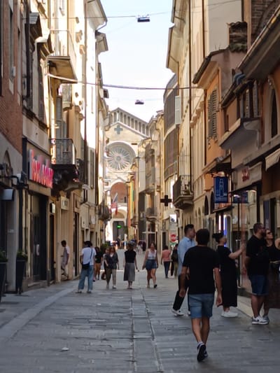 The image depicts a street scene in Piacenza, Italy. The street is lined with multi-story buildings of varying architectural styles, mostly in shades of beige, orange, and brick. The buildings feature windows, balconies, and awnings. Overhead, wires stretch across the street.
A number of people are walking along the stone-paved street. They are dressed in casual summer clothing. The people are in varying positions and are engaged in walking and talking.
In the distance, a church can be seen, identifiable by its large rose window.