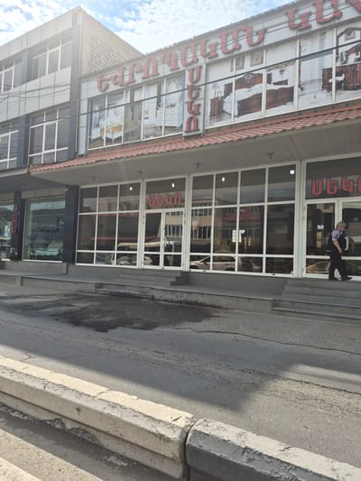 The image shows the exterior of a multi-story building with a storefront at street level. The building's facade features a combination of grey metallic panels and large windows. Above the storefront windows, there is a sign with red Armenian lettering. The storefront itself has large glass windows displaying furniture and interior designs, specifically bedrooms. Red Armenian lettering is also visible above some of the storefront entrances.
A woman in dark clothing, including a vest and pants, is seen walking down a set of concrete stairs to the right of the building's entrance. She is carrying a dark bag or briefcase in her right hand. The street in front of the building is paved and appears wet in some areas, suggesting recent rain or cleaning. A concrete curb with peeling paint runs along the edge of the road. The sky above is partly cloudy. The building is identified as being located in Parakar, Armenia, likely a furniture store.