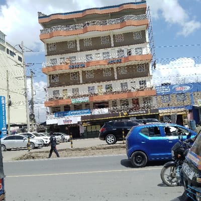 The image captures a street scene in Ongata Rongai ward, Kenya, featuring a multi-story building as its central subject. The building displays a seven-story facade with a distinctive wavy blue roofline and primarily light brown, textured walls. Each floor includes balconies with intricate black and white railings and numerous white-framed windows. Ground floor businesses are identified by signage: "Rongai Clinic," "National Bank," "TriNetra Eye Clinic," and "Venus Medicare Lab Services." A large blue sign on the building's right side advertises "AZAMTV" and "AMAHADI-J UPHOLSTERY & LEATHER SHOP."

In the foreground, a paved asphalt road is present. A blue compact car, likely a Suzuki Alto, is positioned on the right, facing left, notably having a solid black rear wheel. Further down the street, a black SUV is parked. On the left side of the street, a silver sedan and a white sedan are parked in sequence. The front portion of a motorcycle, identified as a Bajaj RE rickshaw, is visible in the extreme bottom right corner.

A single individual, a male in a dark uniform, is depicted walking from left to right on a concrete median strip that separates the parked vehicles from the main road. He carries a small white object in his right hand. To the far left, multiple utility poles with a dense network of overhead wires stand beside a taller, light-colored building. The sky above is partially cloudy, revealing patches of blue.