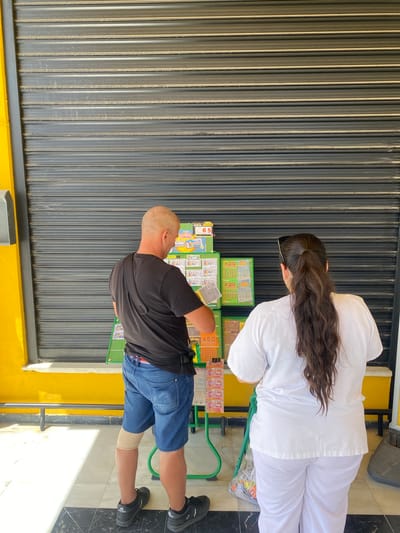 The image shows two people standing in front of a closed metal shutter. The person on the left is a man with a shaved head wearing a black t-shirt and blue denim shorts. He is looking down at something in his hands. He has a compression sleeve on his right leg. The person on the right is a woman wearing a white uniform. She has long, dark hair and is also looking down. They appear to be standing at a stand with lottery tickets and other merchandise. Some of the visible text on the merchandise includes "BOTE DE JACKPOT", "Verano", "0.000€", "ONCE A", "ILLO", "G24538", "ILUSIONATE", "DIO X50", "83520", "SANJUAN", and "x20". The scene suggests it might be daytime, given the lighting, and the setting appears to be an outdoor area, possibly a street or market stall.