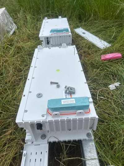 The image shows telecommunications equipment laying in tall green grass outdoors in Weissach im Tal, Germany. The main item is a white, rectangular enclosure, possibly for fiber optic cable connections, with a blue protective cover that reads, "Please tear it off before installation." The enclosure is mounted on a metal bar, with screws and bolts visible. Another, smaller section of the enclosure sits nearby. Scattered around are a pink rectangular object, a piece of white plastic with a honeycomb pattern, and some plastic wrapping.