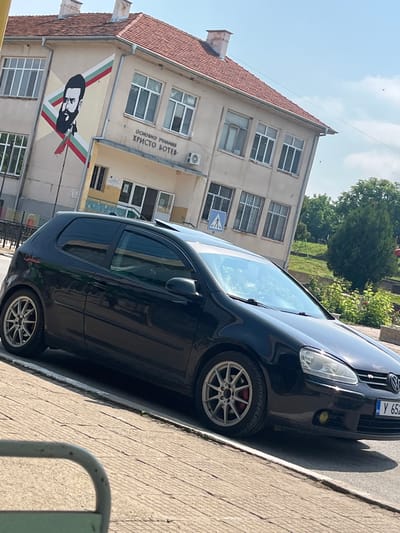 The image, taken with a tilted perspective, depicts a black two-door Volkswagen Golf parked on a paved surface next to a building under clear, sunny skies. Strong sunlight casts distinct shadows, suggesting midday or early afternoon.
The car features multi-spoke silver alloy wheels with visible red brake calipers. Its partially visible Bulgarian license plate, starting with "Y 652", indicates registration in Yambol.
In the background, a two-story beige building with a red-tiled roof is prominently featured. Above the ground floor windows, Cyrillic text identifies it as "ОСНОВНО УЧИЛИЩЕ ХРИСТО БОТЕВ" (Primary School Hristo Botev). Below this text, a large mural displays a stylized black and white portrait of a bearded man, likely Hristo Botev, set against a diagonal backdrop showcasing the white, green, and red colors of the Bulgarian flag. A blue pedestrian warning sign is also visible on the building's facade. Trees and green foliage are present in the background, suggesting an outdoor, possibly suburban or rural, setting.
The scene captures a street in Ботево, Bulgaria, highlighting local architecture and cultural markers such as the Hristo Botev mural and the Bulgarian license plate.