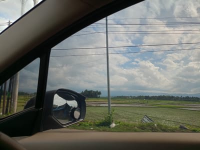 From the interior of a vehicle, the view is of rice fields in Blang Lilue, Indonesia, under a sky with scattered clouds. Utility poles and power lines are visible. The vehicle's side mirror reflects a building. Green vegetation is along the roadside. Mountains are visible in the distance.