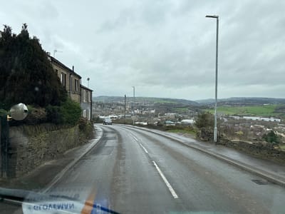 This image depicts a wet, asphalt road descending through an elevated residential area in Calderdale, United Kingdom, under an overcast sky. The viewpoint is from inside a vehicle, evidenced by a windshield reflection displaying reversed text ("WEMARGE" or similar) at the bottom.
On the immediate left, a dry-stone wall runs alongside the road, topped by dense dark green foliage. Behind the foliage, a row of traditional stone-built houses with grey roofs and television aerials is visible. A circular convex safety mirror, mounted on a dark post, projects from the stone wall in the foreground.
The road itself is a two-lane thoroughfare, visibly wet, with a single white broken line marking the center. Multiple darker patches on the asphalt indicate road repairs. A light-colored vehicle, possibly a white car, is observed in the mid-ground, traveling away from the viewer down the curving road.
On the immediate right, another dry-stone wall borders the road, lower than the wall on the left. A tall, slender grey street lamp pole stands vertically alongside the right curb.
In the mid-distance and background, a valley unfolds, revealing a sprawling urban or semi-urban landscape comprised of numerous buildings, including both residential and commercial/industrial structures, nestled amongst rolling green hills. The sky above is uniformly grey and overcast, indicating a cloudy day. No individuals are visible.