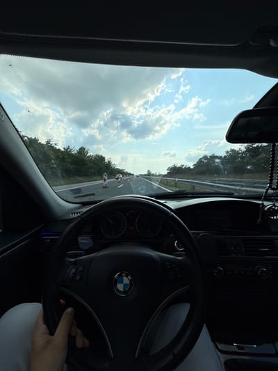 The image provides a first-person perspective from the driver's seat of a BMW car, likely driving on a highway or a multi-lane road in Kaspichan, Bulgaria, during daytime.

Through the windshield, a bright blue sky with scattered white clouds is visible, indicating clear and sunny weather, possibly late morning or afternoon. The road ahead is bordered by dense green trees and foliage. On the right side of the road, orange and white striped traffic barriers or cones suggest ongoing road work or a lane change ahead.

Inside the car, the black leather steering wheel with the distinctive BMW logo is prominent in the foreground. A person's hand is visible holding the lower right portion of the steering wheel. The dashboard and instrument cluster are partially visible behind the steering wheel, showing dials and various buttons. Among the visible controls, "DTC" (Dynamic Traction Control) and what appears to be "AUTO" for climate control can be discerned. A rearview mirror hangs from the top center of the windshield area, from which a string of black beads, possibly prayer beads, is suspended. A phone mount is also attached to the dashboard. The interior appears well-maintained, suggesting an active journey on the open road.