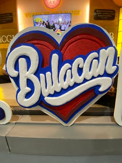 The image shows a large, multi-layered heart-shaped cutout with the word "Bulacan" written in white, 3D lettering with a blue outline. The heart is red with a darker red layer beneath it, and the lettering is outlined in blue and then a darker blue. Behind the cutout, a television screen displays a crowd of people holding up colorful balloons. To the left of the cutout, there is a dark brown plaque with yellow text that appears to be scripture. Above the television, yellow text reads "TO GOD BE GLORY". To the right of the heart cutout, there is another dark brown plaque with yellow text. The cutout is placed on a gray surface, which appears to be a stage or display platform. The overall scene suggests a celebratory or promotional event related to Bulacan. The location in San Jose del Monte, Philippines, is not explicitly visible in the image itself, but the context of "Bulacan" suggests a connection to the province of Bulacan, which is near San Jose del Monte.